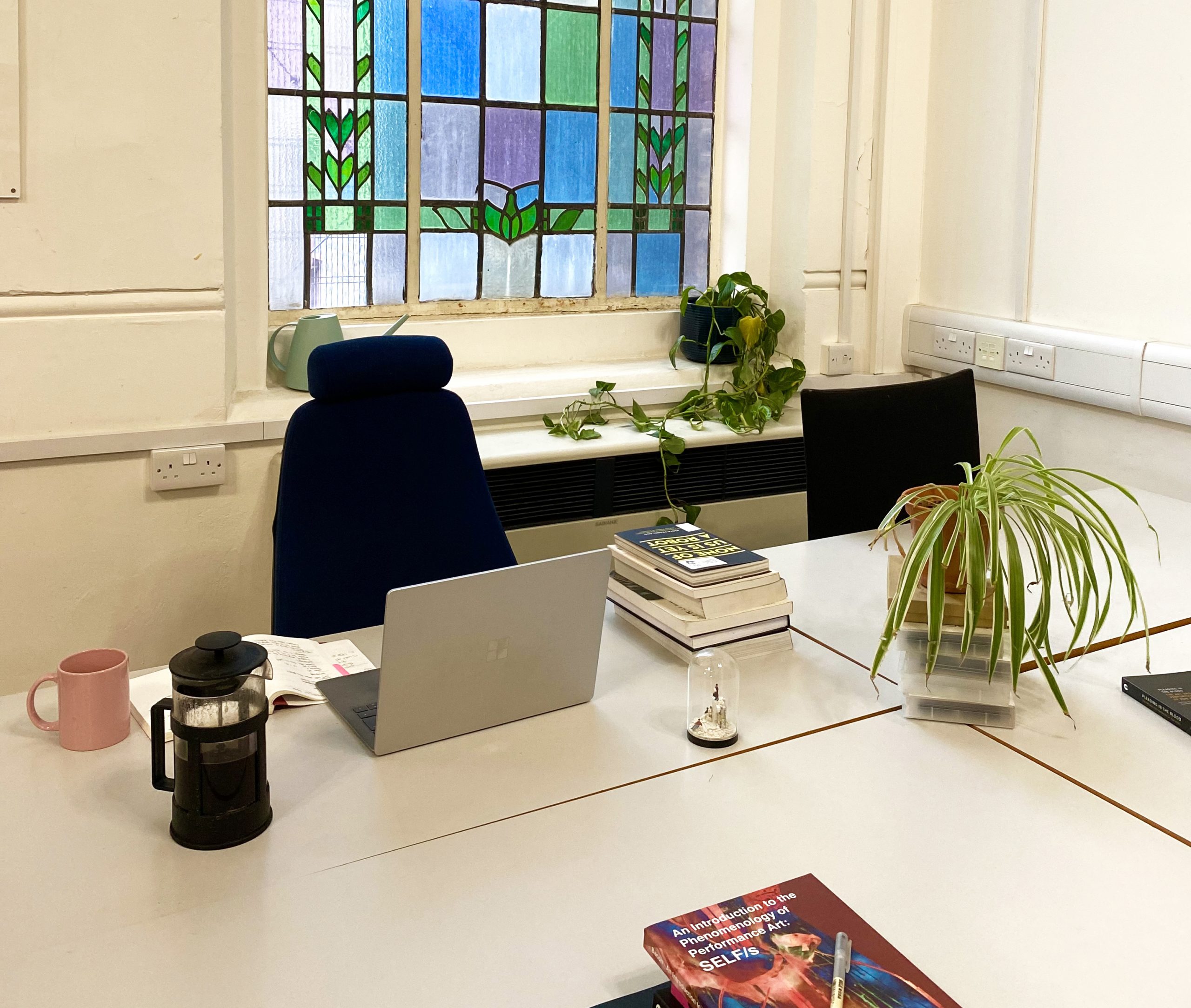 Photo of LADA's Desk Scheme space. Two light grey desks in front of a window. On the desk on the left there is a grey laptop, a pile of books, an open notebook, a French press and a pink mug. There are two plants, one next to it and one behind it.