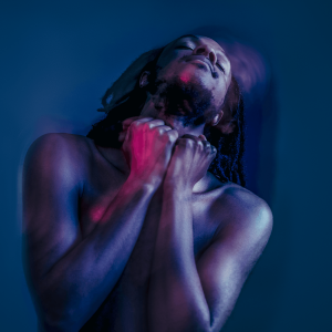Jamal Gerald, who is a Black man with shoulder length dreadlocks, is in a blue-lit room with his head tilted back whilst dancing. A purple light is lighting up his naked body.