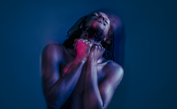 Jamal Gerald, who is a Black man with shoulder length dreadlocks, is in a blue-lit room with his head tilted back whilst dancing. A purple light is lighting up his naked body.