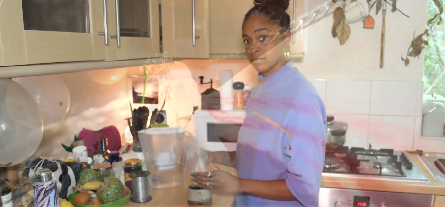 A young light-skinned Black person in a kitchen by the counter. They wear a violet oversized shirt and gold earrings. They look directly at the camera.