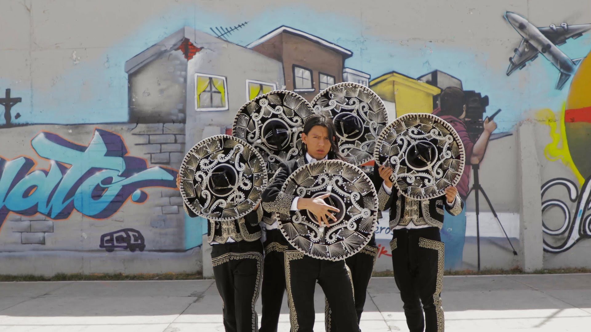 Five mariachis in front of a graffitied concrete wall.