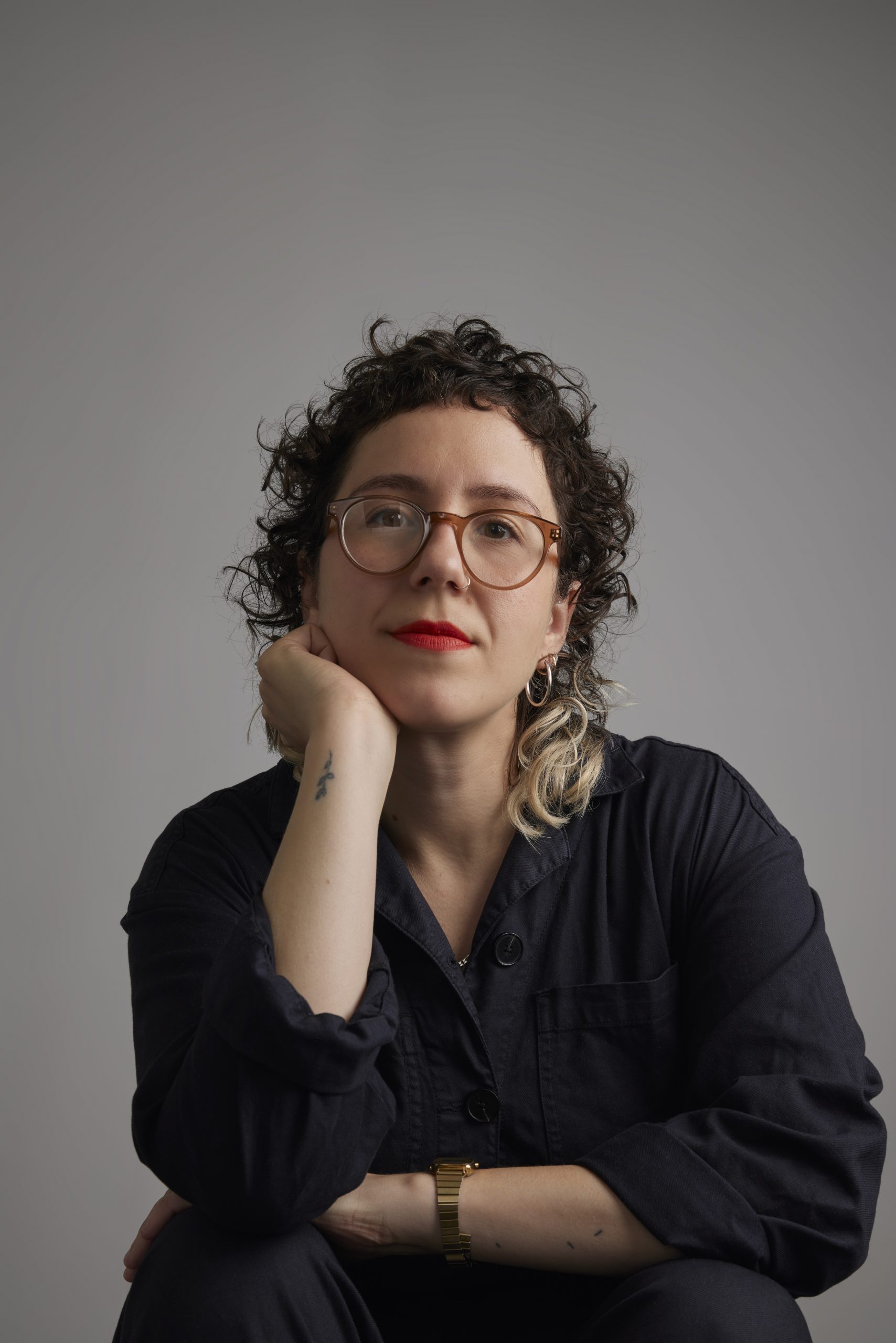Portrait of a white woman in her 30s looking at the camera. She's wearing a black shirt, has curly hair with bleached ends and a short fringe, wears bright red lipstick, light brown glasses, and a gold watch. She has a small tattoo on her wrist.