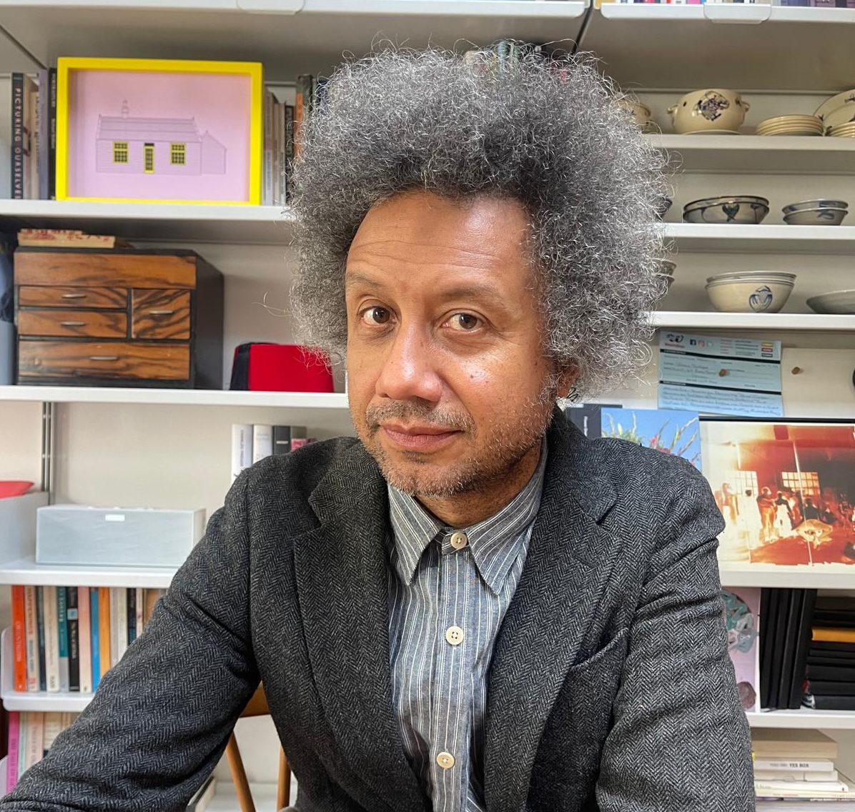 Portrait of a mixed-race man in his 50s. He's wearing a grey blazer and a striped blue and white shirt and has grey afro hair. He's looking at the camera and there's a bookcase covering the wall behind him.