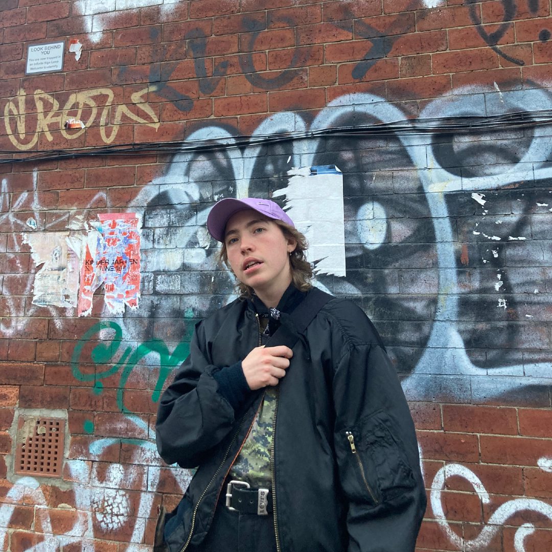 A photo of kane stonestreet in front of a wall with graffities. They're wearing an oversized black bomber jacket and a light purple baseball hat and look at the camera