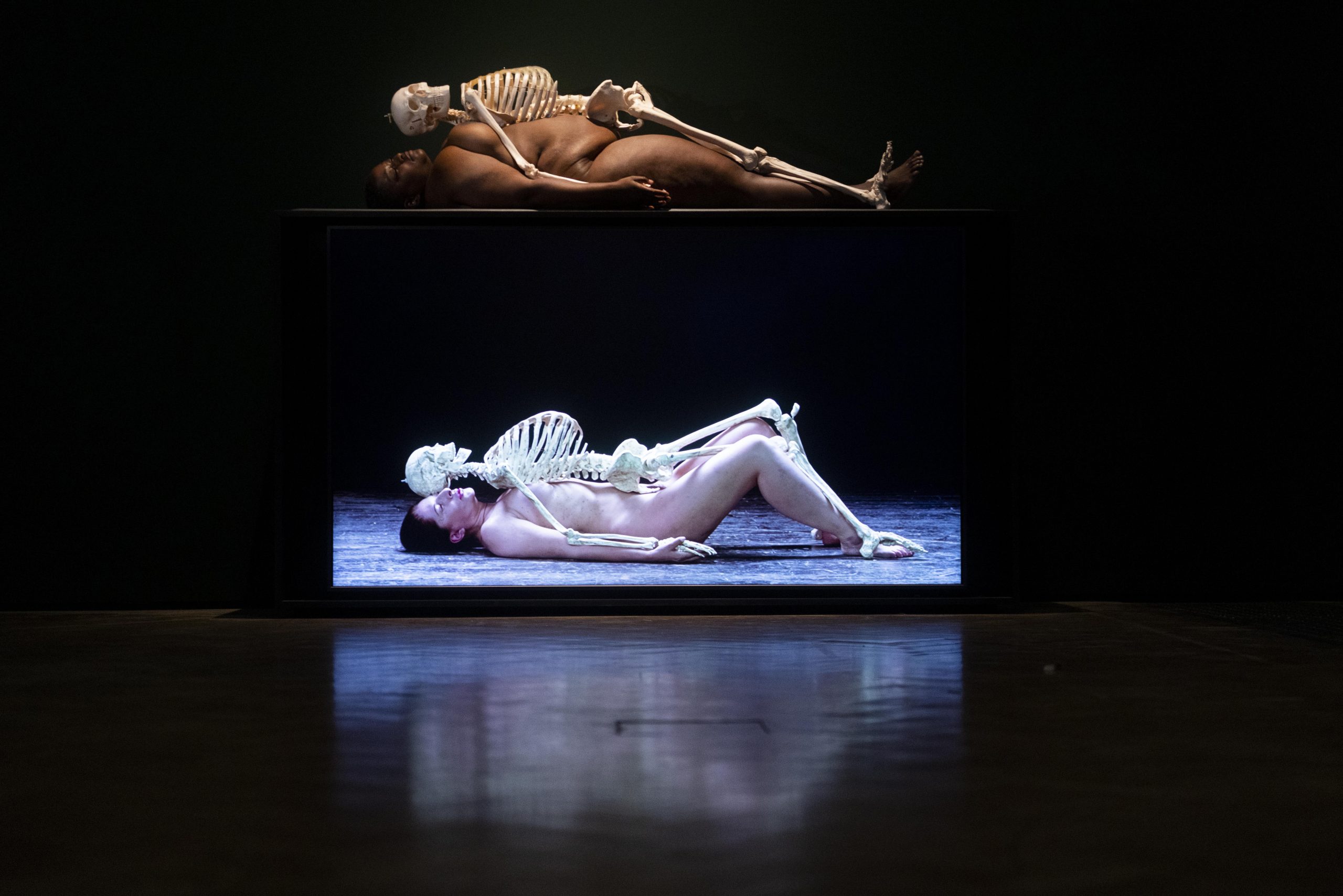 Gallery view of Madinah Farhannah Thompson re-performing 'Nude with Skeleton’ at the Royal Academy of Arts. Madinah is lying on the floor naked, with a human skeleton placed on top of her.