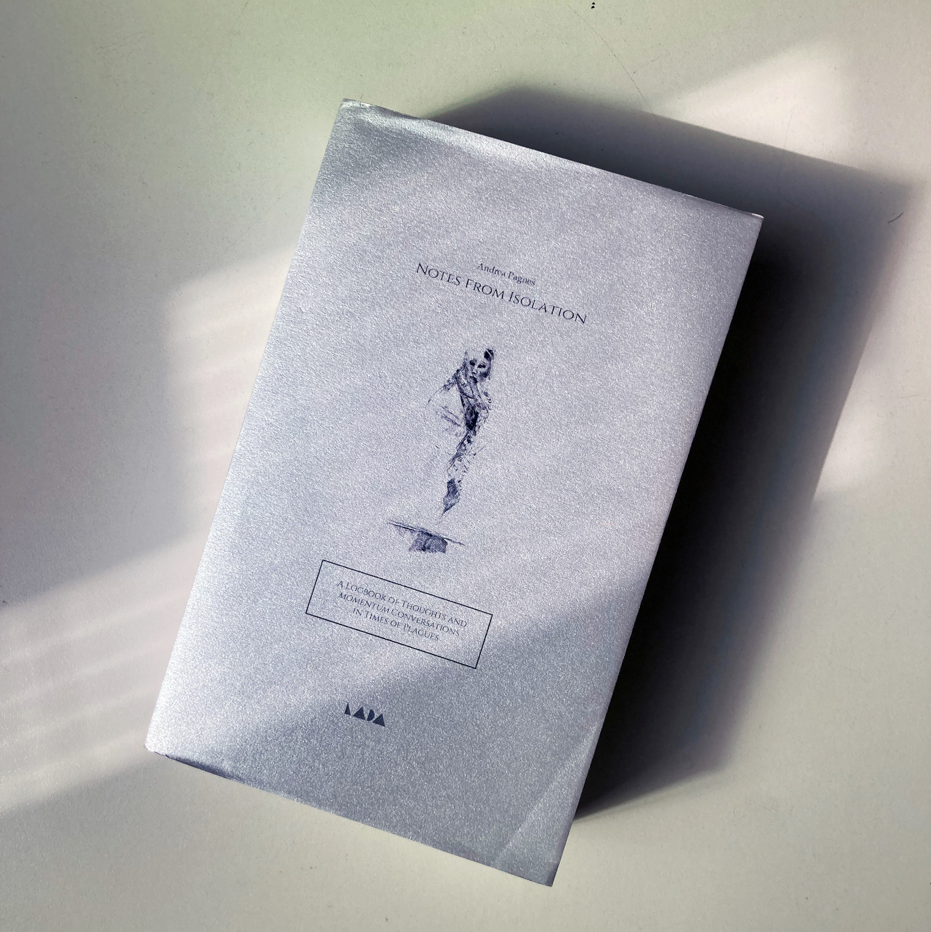 The cover of Notes from isolation on a desk. The cover is silver and features an abstract figure