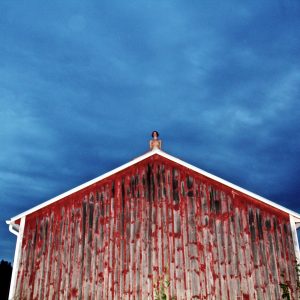 A highly saturated colour photograph of the artist (a white woman) sitting atop a barn, her legs either side of the pitched roof. The camera looks up at her from a dramatically low angle. The sky is dark blue and looming.