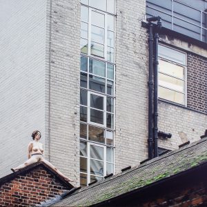 a colour photograph of a woman sitting naked a top a roof in East London. She straddles a pitched roof and sits bolt upright, staring straight ahead. She creates a surreal form amid the grey London buildings.